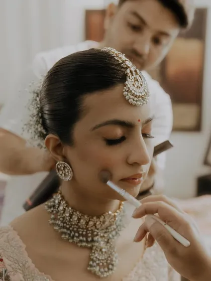 A behind the scenes moment, perfecting a bride's look. The hairstyle is a neat low bun with baby's breath, complementing her intricate jewelry and soft makeup.