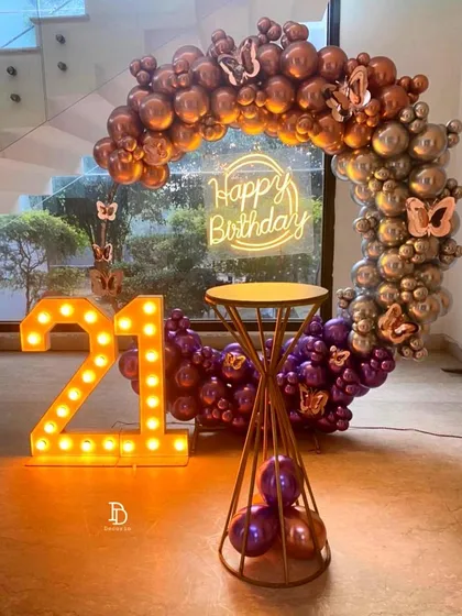 A chic 21st birthday party featuring a circular balloon arch with a mix of metallic rose gold, silver, and purple balloons. The look is completed with large '21' marquee numbers and a 'Happy Birthday' neon sign, creating a perfect photo spot.