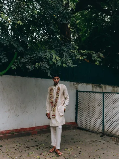 A full-length outdoor shot for Sanchit Sharma. The model stands against a simple white wall, allowing the traditional attire and floral garland to be the main focus.