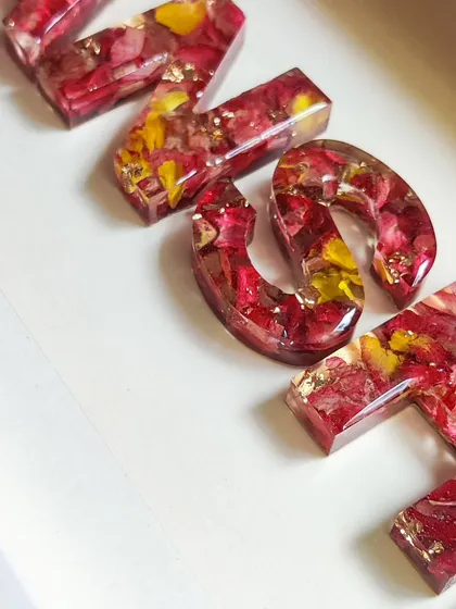 A custom resin name shadow frame is a unique gift for any occasion. Here, the name "ANSHI" is spelled out with letters made from preserved red and yellow flowers, all set in a clean white frame.