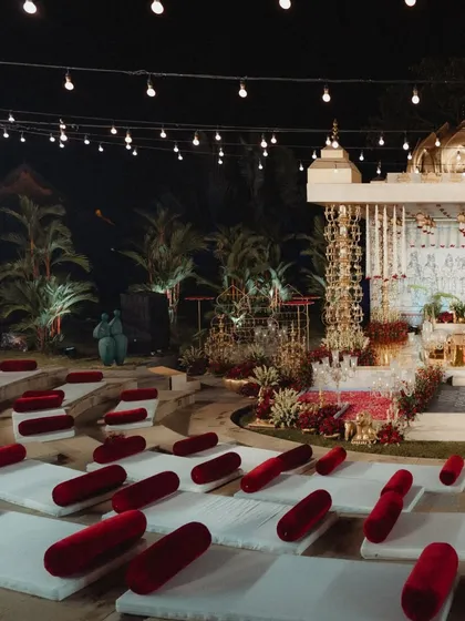 A side view of the amphitheater at night, showing the glowing mandap and the tiered seating dotted with red velvet cushions. The string lights in the background add to the magical, festive atmosphere.