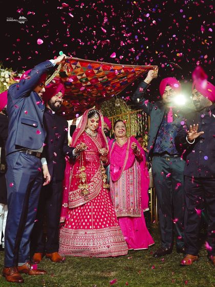 A beautiful bridal entry under a traditional phulkari dupatta, a nod to Punjabi wedding customs. The air is filled with flower petals, creating a vibrant and celebratory atmosphere.