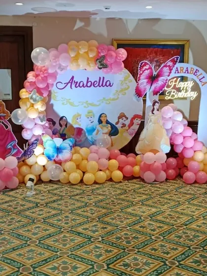A Disney Princess themed birthday party with a custom backdrop. The setup features cutouts of Aurora and Belle, butterfly accents, and a pink and yellow balloon arch.
