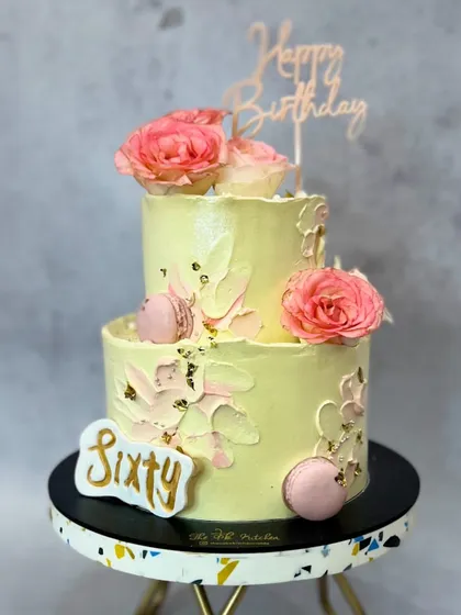 This charming two-tier cake in pastel yellow features a textured buttercream finish, fresh pink roses, and delicate pink macarons. Flecks of gold leaf and a custom 'Sixty' sign make it perfect for a milestone celebration.