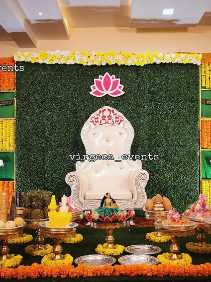 A classic and vibrant setup for a Half Saree ceremony. I use a traditional checkerboard backdrop made with fresh marigold flowers and green fabric, complemented by seer plates, auspicious dolls, and a central lotus motif.