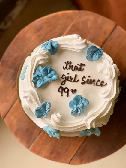 A classic and minimal cake with the message "that girl since 99" and delicate blue buttercream flowers.