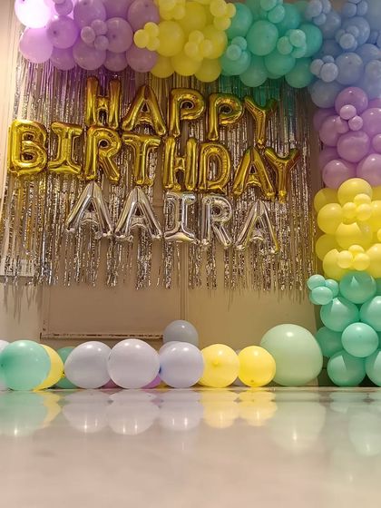 A pastel-themed 3rd birthday decoration for "AAIRA". This low-angle shot shows the balloon arch and foil letters set up against a tinsel curtain in a home.