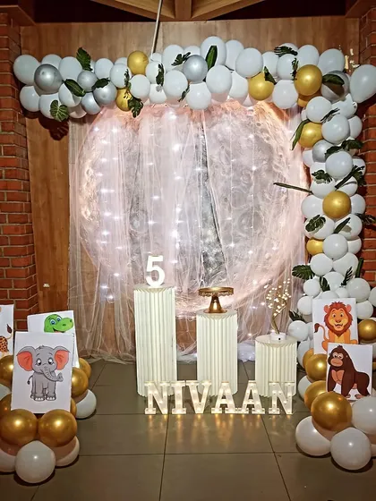 A simple and elegant jungle theme birthday party. This setup uses a white and gold balloon arch with green leaves, animal drawing props, and light-up letters against a brick wall for a modern look.