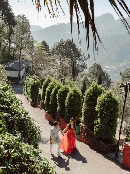 A wide shot of the couple walking down a path lined with cypress trees, capturing the beauty of the hill station.
