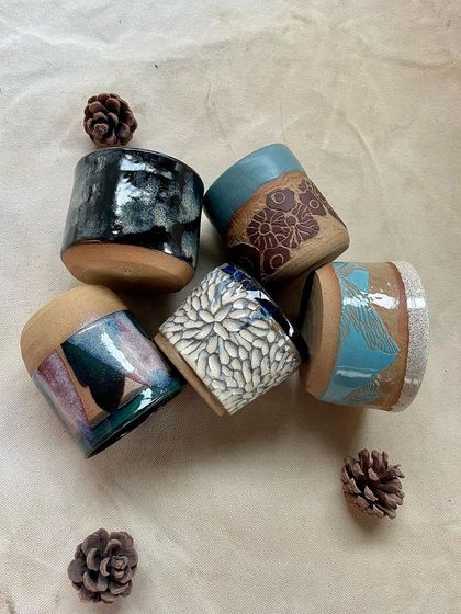 A close-up of five beautifully glazed mugs and cups made by students. The variety of textures and glaze combinations, from floral carvings to abstract drips, is truly inspiring.