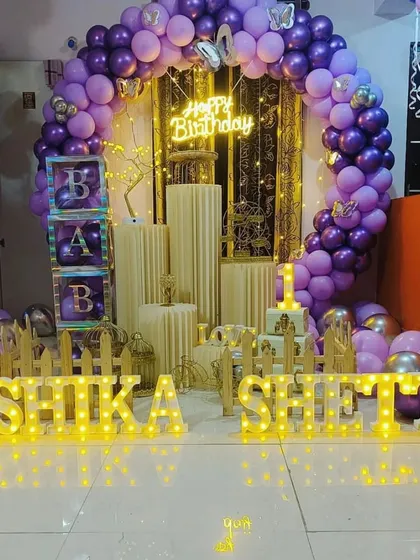 A personalized birthday decoration in purple and gold. This setup features a full balloon arch, a "Happy Birthday" neon sign, and large marquee letters spelling out the name "YASHIKA SHETTY".