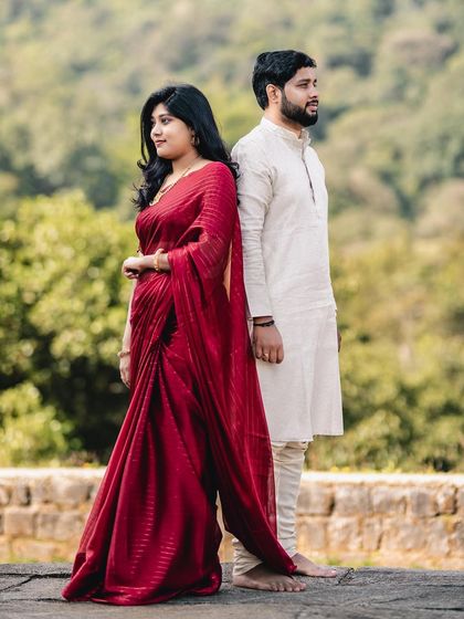 A beautiful, full-length shot of the couple, highlighting the bride-to-be's flowing red saree against the natural green backdrop.