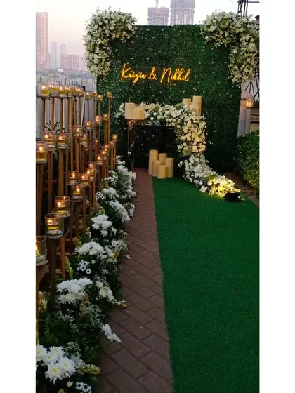 A chic and modern entrance for a special day. A custom neon sign with the couple's names is set into a green hedge wall, while the pathway is lined with flowers and candles for a romantic glow.