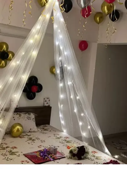 A cozy and romantic bed canopy decorated with fairy lights, with balloons on the ceiling and gifts waiting on the bed.