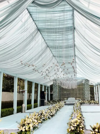 The aisle for the Anand Karaj ceremony, lined with white and blue flowers leading to the main stage. The ceiling is draped in soft, translucent fabric with hundreds of tiny hanging bells.
