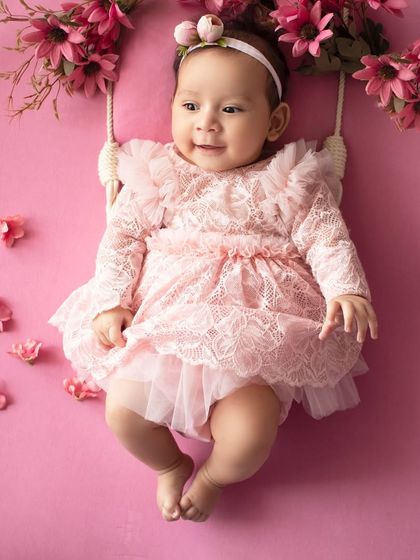 A sweet baby girl in a pink lace dress, smiling from a floral swing prop. A perfect portrait for a little doll.