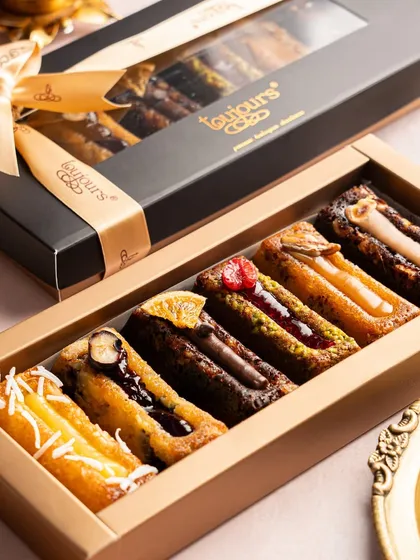 A sophisticated black and gold box filled with an assortment of my signature financier bars. Flavors range from coconut and chocolate chip to pistachio and raspberry, showcasing the variety within a single product line.