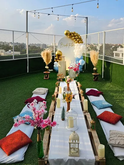 This shows how I can transform a rooftop into a chic boho picnic party. The long, low table is set for guests, complete with colorful cushions, candles, and a stylish backdrop for photos, all under strings of ambient cafe lights.