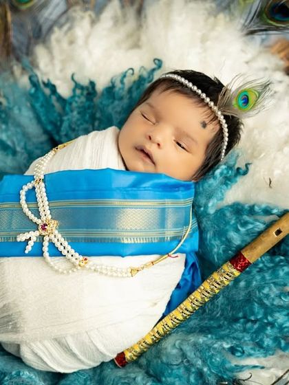 A close-up of the newborn Krishna, wrapped in a traditional silk-bordered cloth and adorned with a pearl necklace and peacock feather.