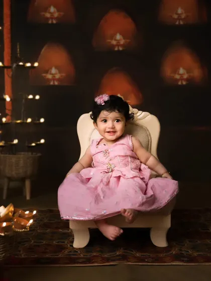 A sweet little girl in a pink dress sits happily in our traditional Diwali setup, surrounded by festive lights.
