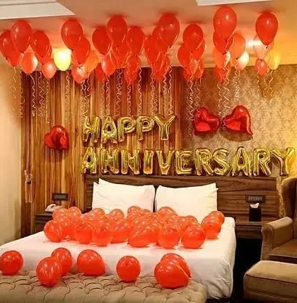 An intimate anniversary room decoration with red balloons, 'Happy Anniversary' foil letters, and balloons on the bed.