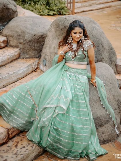 The bride sits gracefully on a rock formation, her flowing green lehenga spread out around her, creating a beautiful contrast with the natural setting.