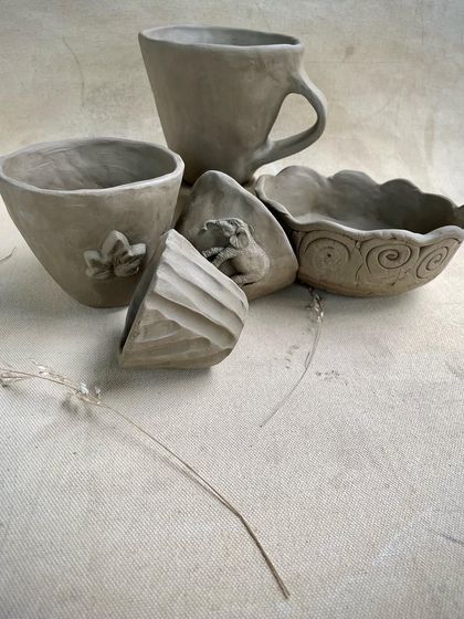 A lovely group of unfired hand-built pieces, including a cup, bowls with scalloped edges, and small pieces with floral and animal motifs. This shows the variety of personal projects students create.