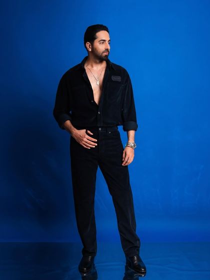 A full-length portrait of Ayushmann Khurrana against a vibrant blue background. The clean, solid color makes the all-black outfit stand out.