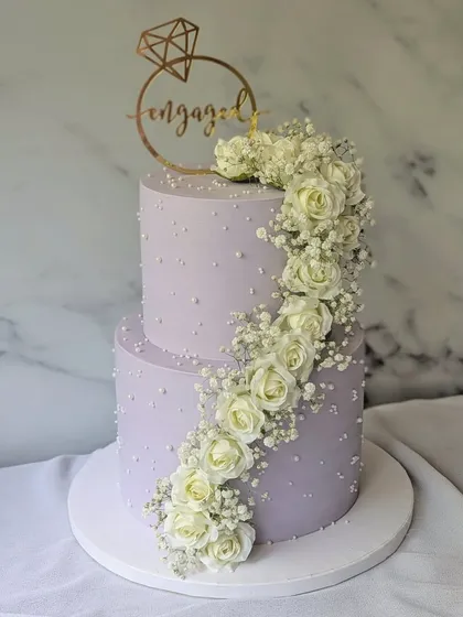 She said yes! This two-tier engagement cake in a lovely lilac shade is decorated with a cascading spiral of fresh white roses and baby's breath. The scattered pearls add a touch of classic elegance.