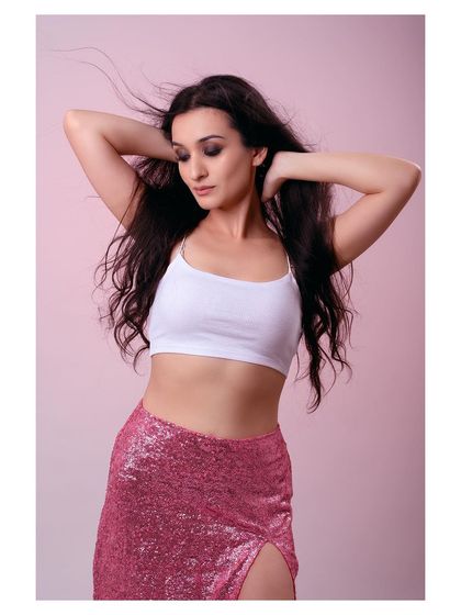 A studio shot with a soft pink background, featuring a model in a white crop top and a pink sequined skirt. The movement in her hair adds a dynamic feel to the image.