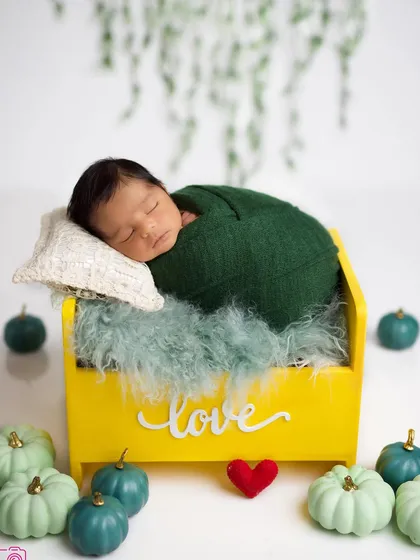 A sweet baby swaddled in green, sleeping in a bright yellow bed surrounded by decorative pumpkins.