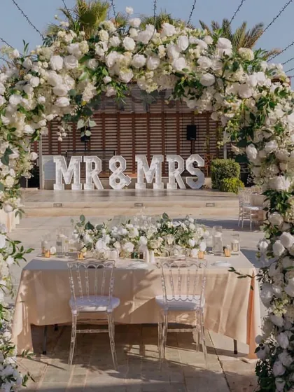 A 'Mr & Mrs' sign and a beautiful white floral arch create a classic and elegant focal point for this outdoor wedding reception.