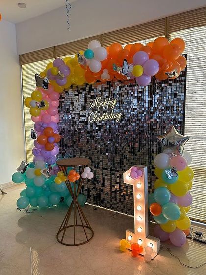 A rainbow butterfly theme against a silver shimmer wall. This colorful balloon arch is perfect for a first birthday, complete with a light-up '1' and a "Happy Birthday" neon sign.
