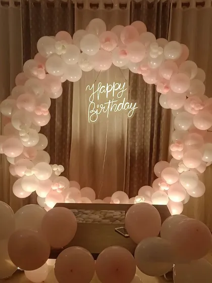 An elegant and minimalist birthday backdrop. This circular arch made of soft pink and white balloons, paired with a "Happy Birthday" neon sign, is perfect for a sophisticated at-home celebration.