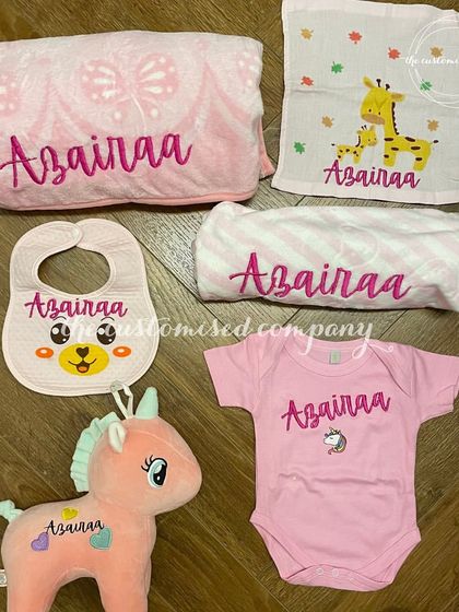 A delightful and comprehensive gift set for a newborn baby girl. It includes a personalized onesie, bib, towels, blanket, and even a soft unicorn toy, all in coordinating shades of pink.