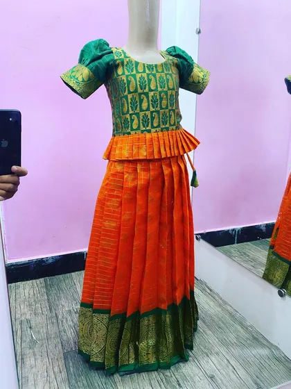 This is a classic kids' Paavada blouse set in a bright orange and green silk combination. The top has a peplum style and puff sleeves, all made with very good stitching for a clean look.