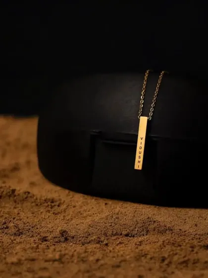 A gold bar pendant with the name 'Vidhi' resting on a textured surface. This product-focused shot highlights the clean font and polished finish.