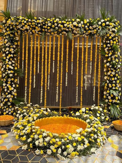 A classic Haldi setup with a twist. The backdrop is framed with yellow and white chrysanthemums, with marigold strings hanging against a dark panel for a beautiful contrast.