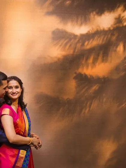 A warm, festive portrait with the shadow of palm trees creating a beautiful pattern on the wall. The couple's embrace and the golden tones make this a perfect holiday season engagement shot.