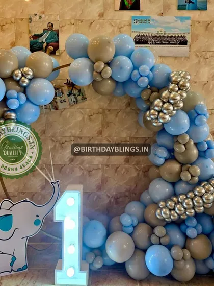 A simple and sweet elephant-themed balloon arch in shades of blue, grey, and silver. The light-up number '1' and the cute elephant cutout holding the balloon string make this perfect for a first birthday.