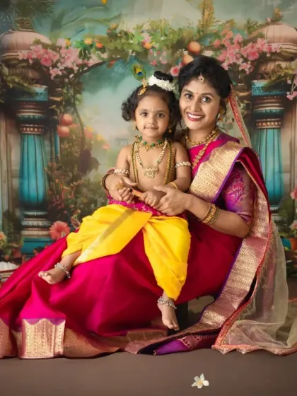 A beautiful portrait of a mother as Yashoda holding her child, dressed as little Krishna. The rich colors of the saree and the backdrop create a regal and warm atmosphere.
