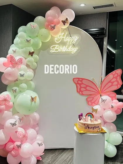 A delicate butterfly-themed birthday setup with a pastel green and pink balloon arch. The white backdrop features a 'Happy Birthday' neon sign and a large pink butterfly cutout.