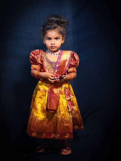A full-length portrait of a little girl dressed in a beautiful pattu pavadai. The dark background and studio lighting emphasize the rich colors and texture of her traditional South Indian dress.