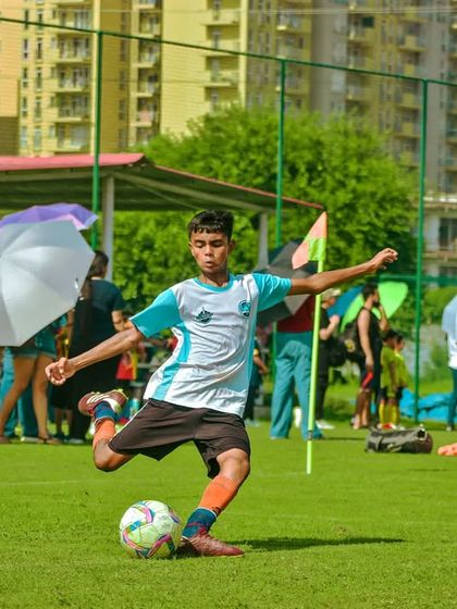 A player takes a powerful kick under the watchful eyes of parents holding umbrellas on a sunny day.