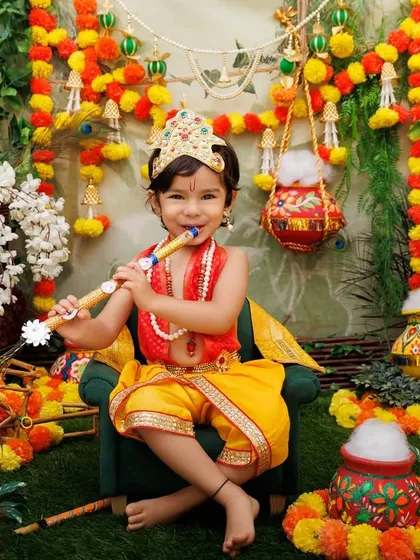 A little Krishna plays the flute in a lush, green setup decorated with marigolds and peacocks.