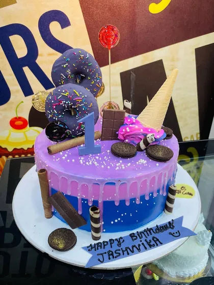 Celebrating a first birthday with a colorful and exciting drip cake. This custom piece combines purple and blue layers and is loaded with fun toppings like sprinkle donuts, an ice cream cone, Oreo cookies, and chocolate wafer rolls.