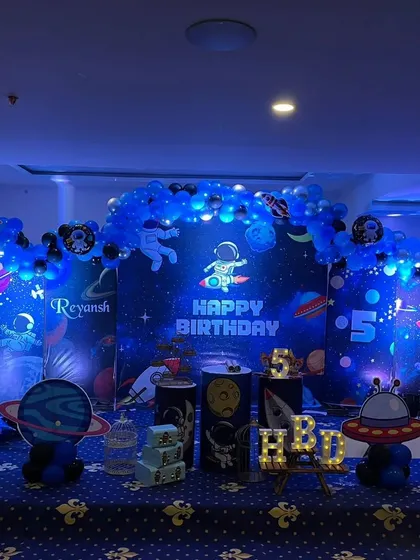 An out-of-this-world outer space theme for a 5th birthday party. I used deep blue lighting to create a galaxy effect, with a star-filled backdrop, astronaut and rocket ship cutouts, planet-themed pedestals, and a light-up number '5'.