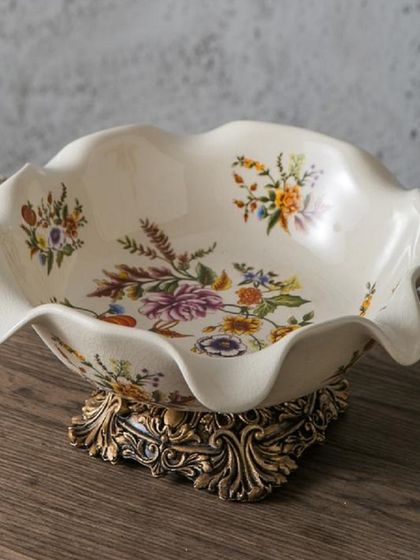 This decorative bowl is a perfect example of Victorian charm, with its scalloped edges, delicate floral paintings, and an ornate, antique-gold finished base. It's ideal for use as a fruit bowl or a standalone centerpiece.