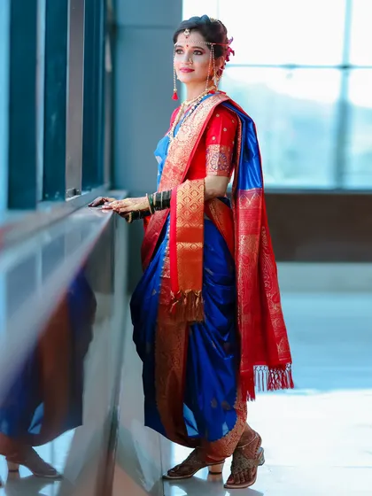 A full-length portrait of a bride in a traditional Maharashtrian Paithani saree. The pose by the window uses natural light to illuminate the rich blue and red silk.