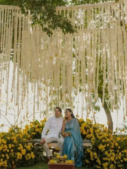 A serene and beautiful Haldi ceremony setting under a tree. The backdrop is a curtain of white and yellow flowers, creating a natural and romantic photo opportunity for the couple.